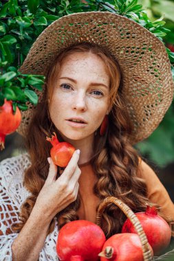 Kırmızı nar bahçesi, turuncu elbiseli ve beyaz şallı mutlu kızıl saçlı kız. Büyük kırmızı meyveli kadın portresi