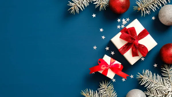 Kırmızı kurdeleli beyaz hediye kutuları, balo süslemeleri ve koyu mavi arka planda konfetili çam ağacı dalları. Noel hediyesi konsepti. Düz yatış, üst görünüm.