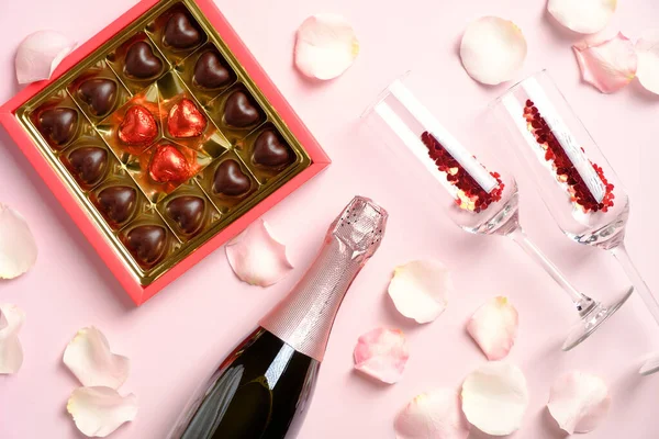 Romantic dinner. Box of chocolates, bottle of champagne and glasses with confetti on pink background decorated with rose petals. Valentines Day celebration concept.