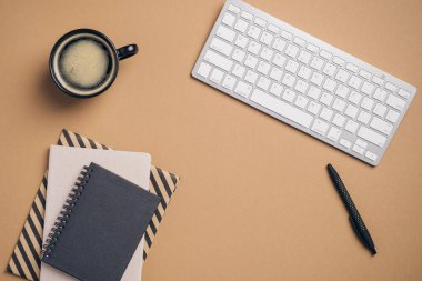 Düz uzanın, üst kattaki ofis masasında klavye, bir fincan kahve, kağıt not defterleri, kahverengi arka planda bir kalem. Modern işyeri konsepti