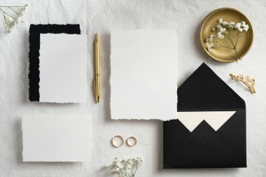Düğün kırtasiyesi hala hayat sahnesinin üst görüntüsü. Düz davetiye kartları şablonları, siyah zarflar, altın kalem, yüzükler, tekstil arka planında çiçekler. Kopyalama alanı olan üst görünüm.