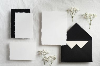 Pastel beyaz tekstil arka planında kadın düğün kırtasiyesi modeli. Çingene çiçekleri, boş kâğıt kartları, siyah zarf üst görünümü.