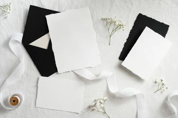 Black envelopes and white paper cards with flowers and ribbon on textile background. Flat lay, top view. Wedding flat lay composition.