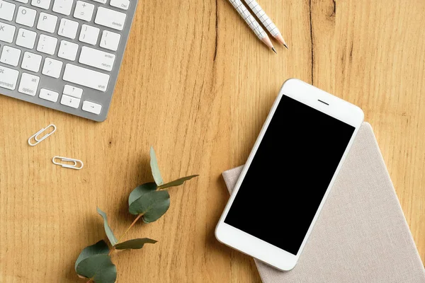 Modern office desk top view. Flat lay smartphone with empty screen mockup, pc keyboard, eucalyptus leaf, stationery on wooden table