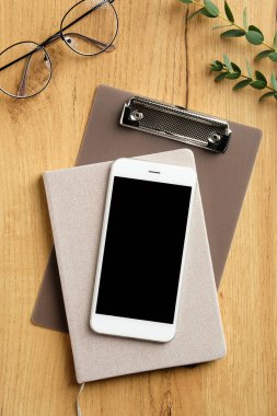 Akıllı telefonlu ahşap ofis masası, kağıt defter, dosya, kadın gözlüğü, okaliptüs yaprakları. Üst görünüm, düz görünüm, kopyalama alanı.
