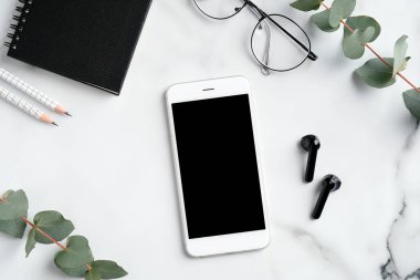 Akıllı telefon modeli, kablosuz kulaklık, gözlük, not defteri, mermer arka planda okaliptüs yaprakları olan kadınsı çalışma alanı. Düz konum, üst görünüm, kopyalama alanı.