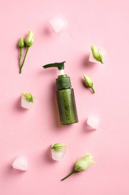 Yeşil duş jeli şişesi, buz küpleri ve pembe arka planda çiçekler. Düz yatış, üst görünüm.