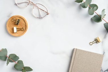 Üzerinde defter, gözlük, okaliptüs dalları olan mermer ofis masası. Zarif, kırsal, kadınsı çalışma alanı. Kopyalama alanı olan üst görünüm, düz yatay.