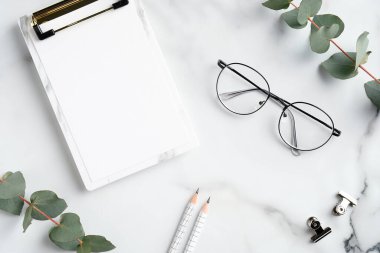 Kağıt klasörlü, gözlüklü, mermer arka planda okaliptüs dallı zarif bir ofis masası. Asgari tarz çalışma alanı. Düz yatış, üst görünüm.