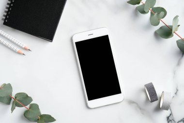 Düz telefon modeli, kağıt defter, kalemler, mermer arka planda okaliptüs yaprakları. Üst düzey ofis masası, zarif kadınsı çalışma alanı..