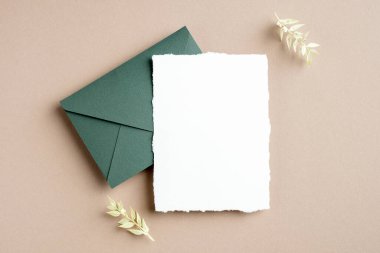 Rustik düğün davetiyesi maketi, yeşil zarf ve pastel bej arka planda kurumuş çiçekler. Düz konum, üst görünüm, kopyalama alanı.