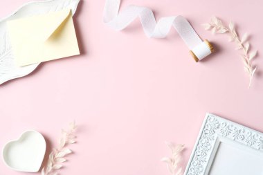 Resim çerçeveli, kurdeleli, çiçekli, pembe arka planda zarfı olan zarif bir kadın çalışma alanı. Düz yatış, üst görünüm.