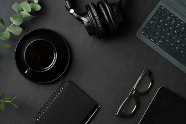 Laptop bilgisayarı, kahve fincanı, kulaklık, gözlük, kağıt defter, siyah masa tableti ile modern ofis çalışma alanının en üst görünümü.