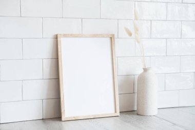 Boş dikey ahşap çerçeve maketi ve masadaki vazoda kurumuş çiçekler. İskandinav iç mimarisi, asgari, zarif. Beyaz karo duvar arkaplanı.