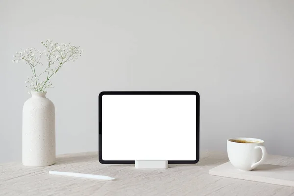 Tablet screen mockup, vase of flowers and coffee cup. White wall on background. Cozy, hygge, nordic style modern workspace