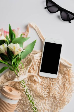 String bag with cup of coffee, flowers, smartphone mockup on white background. Elegant feminine accessories. Sustainable lifestyle concept.