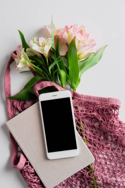 Bahar çiçeklerinin olduğu çevre dostu bir alışveriş çantası, akıllı telefon modeli, beyaz masanın üzerinde bir defter. Düz yatış, üst görünüm.