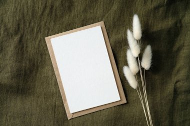 Yeşil tekstil arka planında kurumuş çiçekler olan beyaz davetiye modeli. Düz yerde, üst tarafta. Düğün davetiyesi şablonu