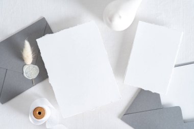 Düğün kırtasiyesinin üst görüntüsü. Düz beyaz davetiye kartı, gri zarflar, kurumuş çiçekler, beyaz arka planda kurdele..