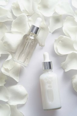 Beyaz arka planda gül yaprakları olan gerekli yağ şişesi ve serum. Düz yatış, üst görünüm