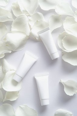 Beyaz arka planda gül yaprakları olan kozmetik tüpleri. Düz yatış, üst görünüm.