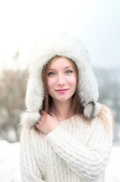 Retrato de uma jovem menina bonita em um boné com abas de ouvido em um parque de inverno fresco — Fotografia de Stock