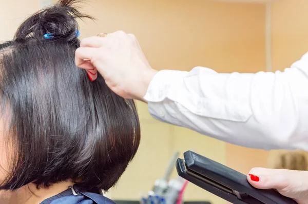 The hairdresser straightens the hair of a young brunette with a Curling iron and combs her hair with a comb.