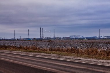 Sonbaharın sonlarında sanayi manzarası. Endüstriyel bir fabrikanın bacalarını tüttürmek, bulutlu bir sonbahar gökyüzünün altında karla kaplı tarlalar.