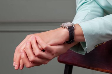 Sandalyenin arkasına yaslanmış turkuaz ceketli bir kadın. İki kadın eli ve bir saat birbirine bağlanmış. Yakından.