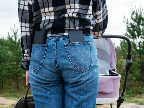 A young mother walks with a stroller in nature and charges her phone with a power Bank. The use of gadgets in life. The concept of a modern mom on a walk. powerBank and phone in your jeans pocket.