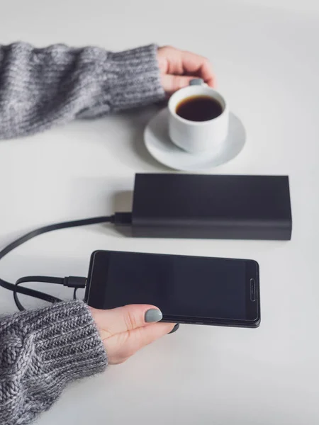 Power bank and phone are black. Power bank charges the phone on the table. Smartphone and a Cup of coffee in women\'s hands.