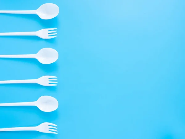 Disposable tableware, white plastic forks and spoons isolated on a blue background with a copy of the space. Modern problems of plastic product recycling and recycling. Environmental pollution.