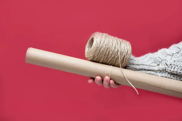 Hand holding a roll of Kraft paper and a reel of rope on a red background, close-up. Gift wrapping with your own hands in a retro style concept.