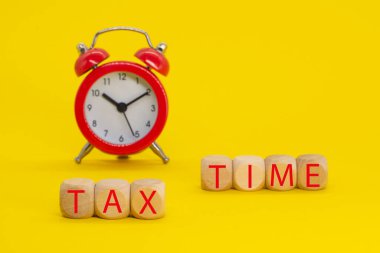 Tahta küplere yazılmış vergi zamanı ve sarı arka planda çalar saat. Vergi ödeme hatırlatması veya yıllık vergilendirme kavramı.