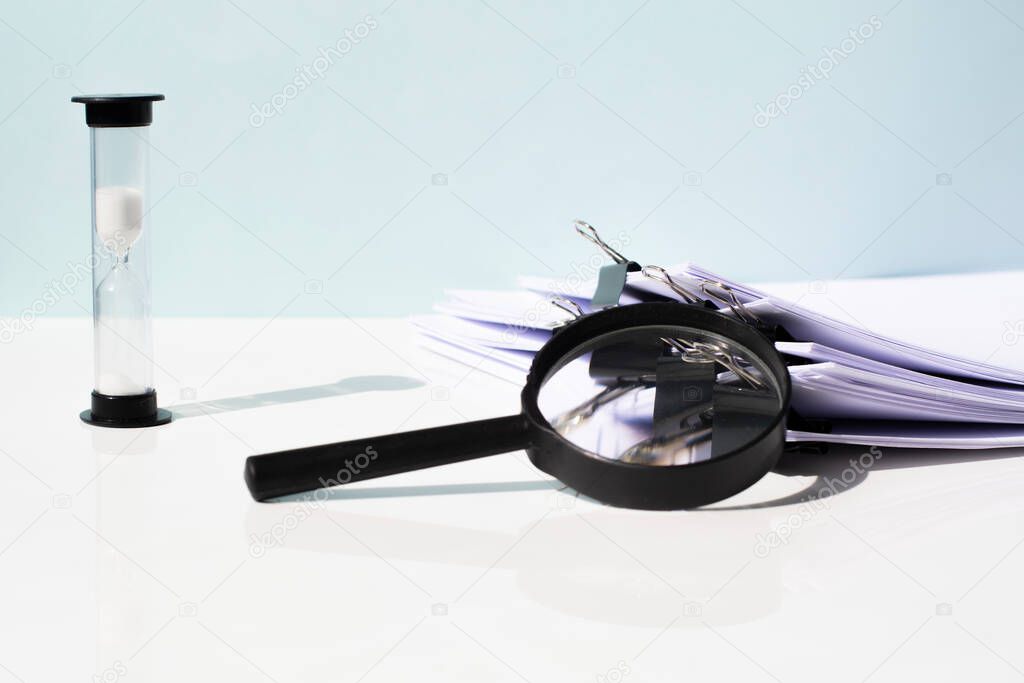 Scanning business documents. Magnifying glass on the stack of documents and an hourglass on a white table