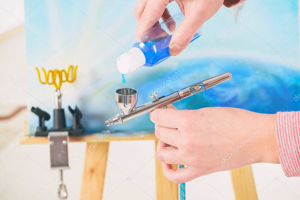 Artist fills the airbrush canister with blue paint. An easel with a painting is visible in the background