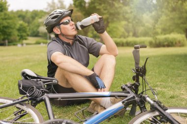 Bisikletçi çayırda dinlenirken şişeden su içer.