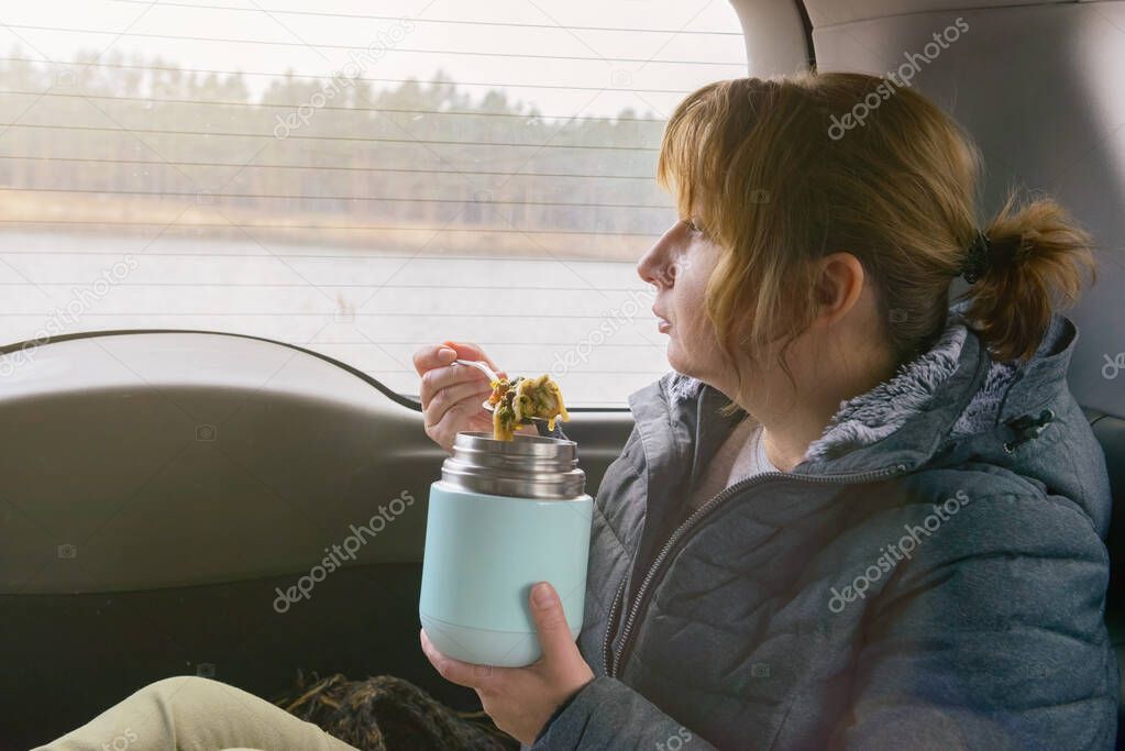 Woman sits in her car and looks out the window at the lake while eating a hot meal from a thermos. The concept of eating food outside and having a picnic