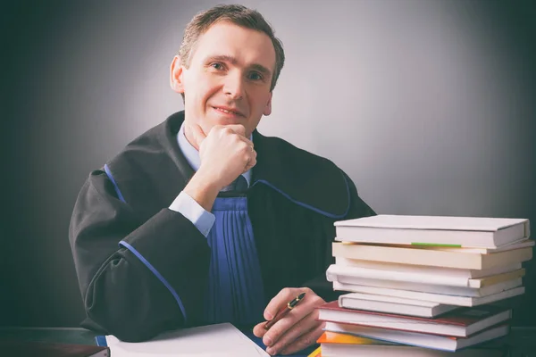 Stock image Smiling lawyer in a gown sits at a desk full of books against a dark background