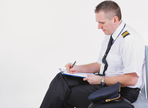 Airline captain pilot fills out the paperwork before the flight. A cap and a suitcase are lying next to him.