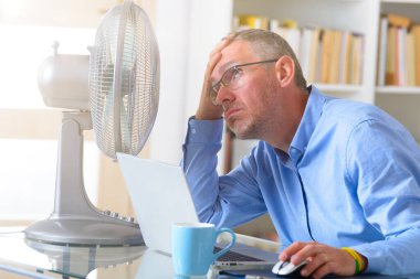 Adam ofiste çalışırken sıcaktan muzdarip oluyor ve vantilatörün karşısında serinlemeye çalışıyor.