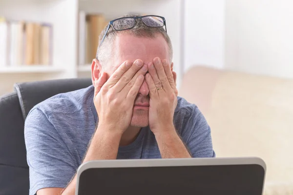 疲れたフリーランスの男はノートパソコンで作業しながら彼の目をこすり — ストック写真