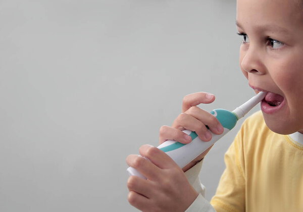 Маленький мальчик чистит зубы электрической зубной щеткой. Изображение с серого фона.