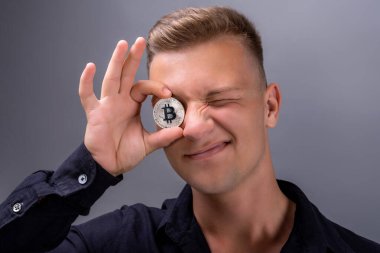 Genç adam gri arka planda bitcoin tutuyor. Gülümsüyor ve göz kırpıyor. Elinde gözlerinin yanında bir bozuk para tutuyor. Kripto para birimi pazarı büyüyor..
