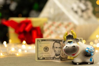 Üzerinde 5 dolarlık banknot olan şirin gri inek heykelciği, parlak ışıklı Noel hediyeleri ve bulanık arka planda xmas ağacı, 2021 yılının boğa sembolü, bayram tebrik kartı, seçici odaklı.