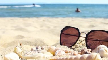 Deniz kabukları ve güneş gözlükleri yaz kumsalında, bulanık insanlar ve arka planda turkuaz deniz jetsileri, fotokopi alanı düşük açı, ön seçici odak, tatil beldesi konseptini gevşetin