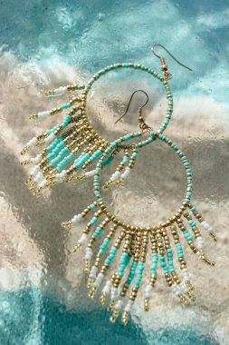 Turkuaz boho mücevher küpeleri cam üzerinde taze su püskürtüsü ve mavi kumlu bej arka plan plaj tatil tarzında yaz aksesuarı tropikal deniz mücevherleri, dikey görüntü