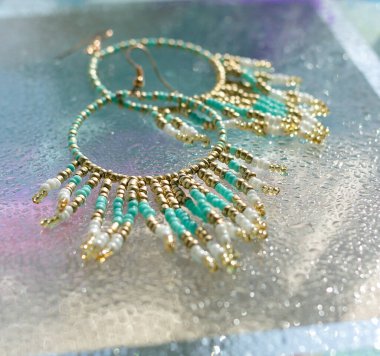 Altında parlak cam ve mavi arka plan bulunan turkuaz boho küpeler, plaj tatili tarzında yaz aksesuarları, tropik deniz mücevherleri konsepti, düşük açılı parlak görüntü.