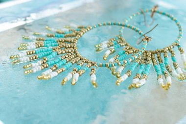 Altında su ve mavi kumlu bej arkaplan bulunan turkuaz boho mücevher küpeleri, plaj tatili tarzında yaz aksesuarı, tropik deniz mücevheri konsepti, alçak açılı görüntü.