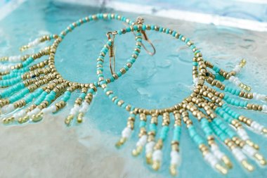 Altında su ve mavi kumlu bej arkaplan bulunan turkuaz boho mücevher küpeleri, plaj tatili tarzında yaz aksesuarı, tropik deniz mücevheri konsepti, alçak açılı görüntü.